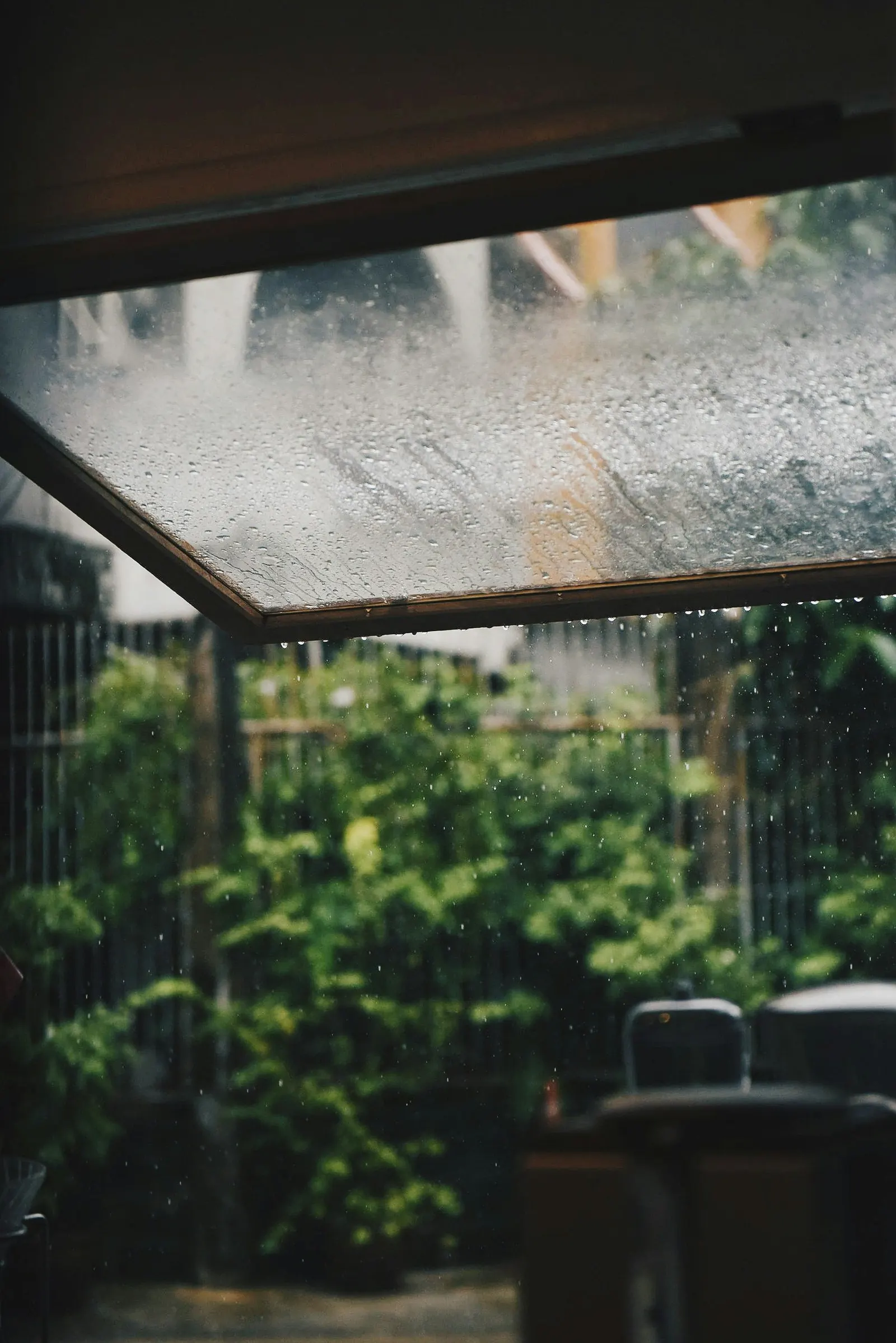 從棚下往外看，框住的玻璃採光罩上布滿雨滴，雨水從邊緣滴落，下方是模糊的綠色庭院與椅子
